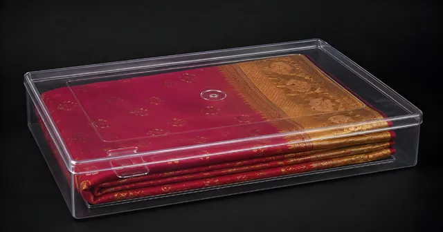 A crystal-clear saree box holding a folded magenta saree with a gold zari border, photographed on a dark background.